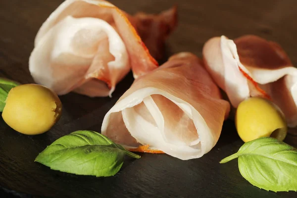 Curled Slices of Delicious Prosciutto with basil  leaves and olives on granite board. — Stock Photo, Image