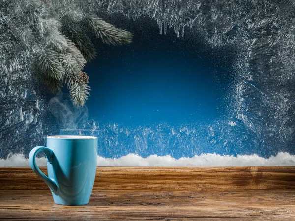 Kubek gorącej herbaty, mrożone okna i choinki przeciwko niemu. — Zdjęcie stockowe