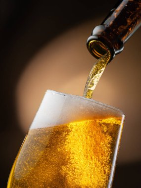 Şişeden çıkan biralar bira bardağına dökülüyor, bu da bir sürü köpüğe ve köpüğe neden oluyor. Koyu kahverengi arkaplan.