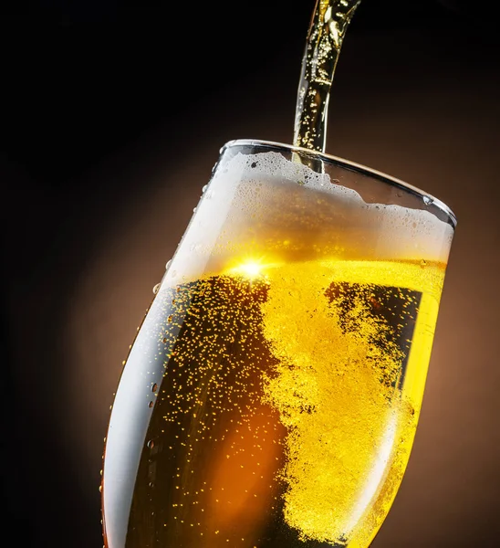 Jet of beer is poured into a beer glass, causing a lot of bubbles and foam. Dark brown background.