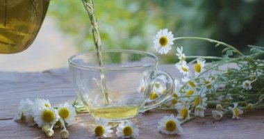 Papatya çayı şeffaf bir çaydanlıktan cam bir fincana dökülür. Eylem, yaz aylarında kırsal bir terasta güneş ışınları eşliğinde gerçekleşiyor. Yakınlarda bir ilaç papatyasının çiçekleri var..