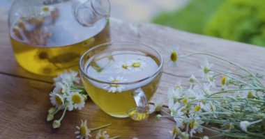 Cam çaydanlık ve fincanda papatya çayı, kamera hareket ediyor. Eylem, yaz aylarında kırsal bir terasta güneş ışınları eşliğinde gerçekleşiyor. Yakınlarda bir ilaç papatyasının çiçekleri var..    