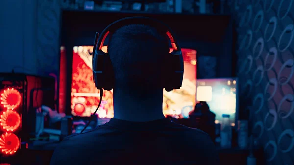 a gamer is sitting in front of a large monitor with headphones,a silhouette in headphones against the background of a monitor with a computer game.photo taken in a dark key in neon light. guy plays computer games at home