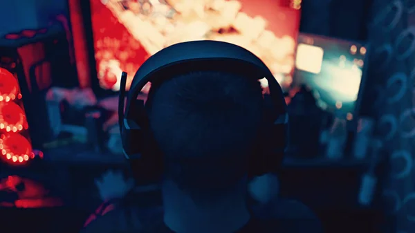 a gamer is sitting in front of a large monitor with headphones,a silhouette in headphones against the background of a monitor with a computer game.photo taken in a dark key in neon light. guy plays computer games at home