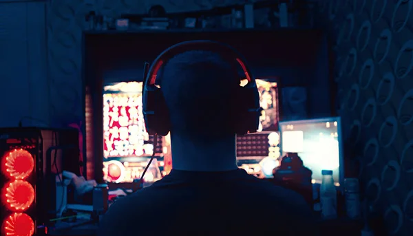 a gamer is sitting in front of a large monitor with headphones,a silhouette in headphones against the background of a monitor with a computer game.photo taken in a dark key in neon light. guy plays computer games at home