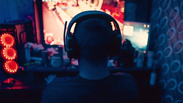 一个游戏玩家坐在一个带耳机的大型监视器前 耳机中的轮廓在一个带着电脑游戏的监视器的背景下 那家伙在家里玩电脑游戏 — 图库照片