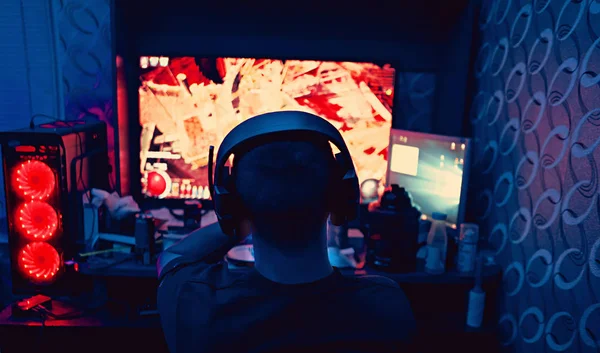 a gamer is sitting in front of a large monitor with headphones,a silhouette in headphones against the background of a monitor with a computer game.photo taken in a dark key in neon light. guy plays computer games at home