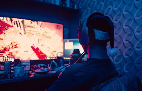 a gamer is sitting in front of a large monitor with headphones,a silhouette in headphones against the background of a monitor with a computer game.photo taken in a dark key in neon light. guy plays computer games at home