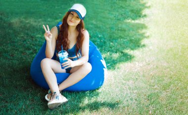 Kafkas görünümlü genç bir kadın çimenlikte bir puf üzerinde oturuyor ve eğleniyor, duygular besliyor, mavi spor giyim ve şapka, yaz tatili konsepti