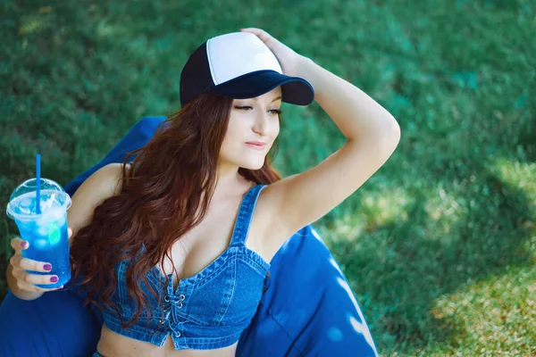young woman of Caucasian appearance sitting on a puff on a lawn and having fun, makes emotions, she is in blue sportswear and a cap, summer vacation concept