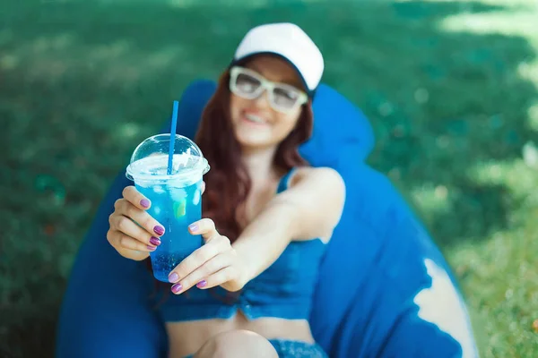 young woman of Caucasian appearance sitting on a puff on a lawn and having fun, makes emotions, she is in blue sportswear and a cap, summer vacation concept