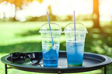 Yazın dışarıda 2 kokteyl, güneş gözlüklerinin yanındaki tepside, çimlerin etrafında, mavi gölde, güneş parlaması.