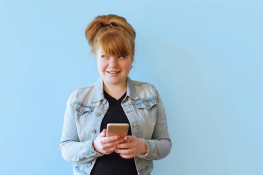 Kafkasyalı neşeli bir kız cep telefonuyla poz veriyor, modern bir akıllı telefona mesaj yazıyor, boş zamanlarında komik videolar izliyor, iyi bir internet bağlantısından hoşlanıyor, mavi izole edilmiş bir geçmişi var.