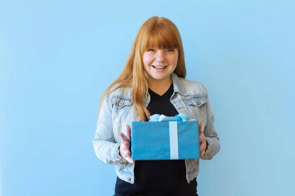 a happy woman received a blue gift box with a bow. She is happy and flattered by attention. Girl dancing on a blue background. she is wearing a denim jacket