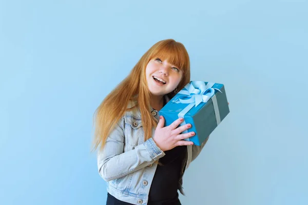 a happy woman received a blue gift box with a bow. She is happy and flattered by attention. Girl dancing on a blue background. she is wearing a denim jacket
