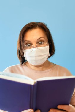 Koruyucu antiviral maskeli olgun bir kadın izolasyon kitabında evde oturuyor ve mavi bir arka plan karantinasında kitap okuyor..