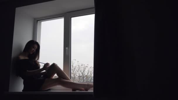 Silhueta de menina atraente sentado no peitoril da janela — Vídeo de Stock