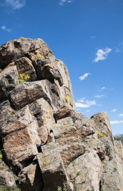 kaya üzerinde taşların yakın çekim görünümü, arka planda mavi gökyüzü, kayalar arasında sarı çiçekler, basamaklı taşlar, dikey yönelim