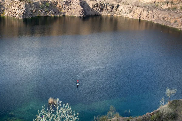 男は青い湖、トラックは葉の真ん中に sup のカヤックします。岩の海岸 — ストック写真