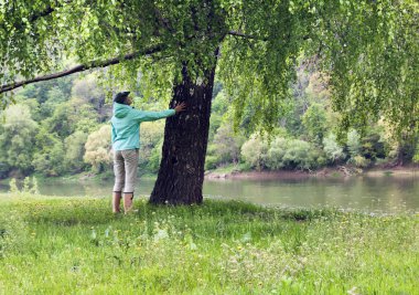 Kız şapka ve lastik Çizmeli Nehri yakınında büyük ağacın altında duruyor. Kadın dokunuşlar huş ağacı