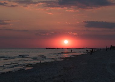 Sahilde kırmızı günbatımı. İnsanlar siluetleri ve martı sahilde. İskelede manzarası