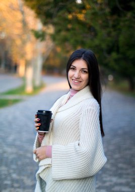 Sonbahar parkında beyaz hırkalı genç bir kadın, elinde siyah bir fincan kahveyle.
