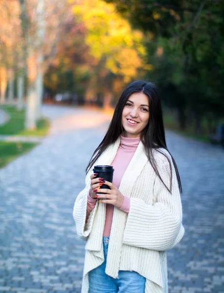 young female in white cardigan in the autumn park with black cup of coffee