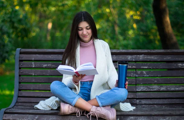 Молодая женщина читает книгу на скамейке в парке, термокружка на скамейке — стоковое фото