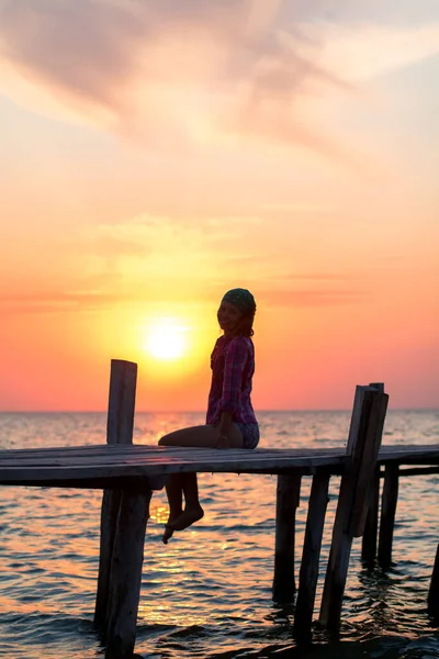 girl sitting dangling legs on the old wooden pier and admires of red sunset in the sea