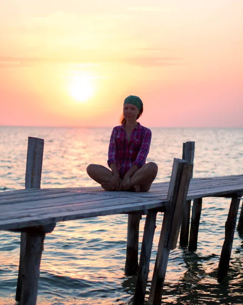 Meisje zit in lotus poseren op een oude houten pier en geniet van de zonsondergang op de zee — Stockfoto