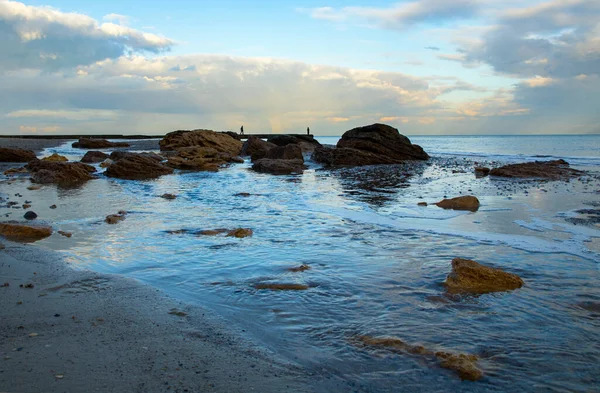 Λείο μπλε νερό και πέτρες στην παραλία, δύο αγνώριστη σιλουέτα στην προβλήτα — Φωτογραφία Αρχείου
