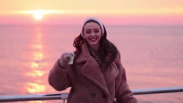 海の遊歩道でコーヒーを飲みながら毛皮のコートで笑顔の若い女の子は美しい日の出を見るのが嬉しい — ストック動画