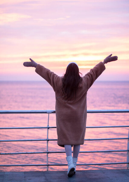 A young girl in a fur coat with a hot drink welcomes a red sunrise on the sea promenade