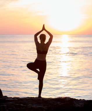 Kızın kumsalda büyük taşların arasında yoga eğitimi var. Denizde kayalar. gün doğumu