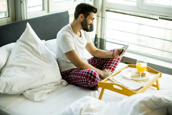 Joven desayunando en la cama y usando un teléfono móvil — Foto de Stock