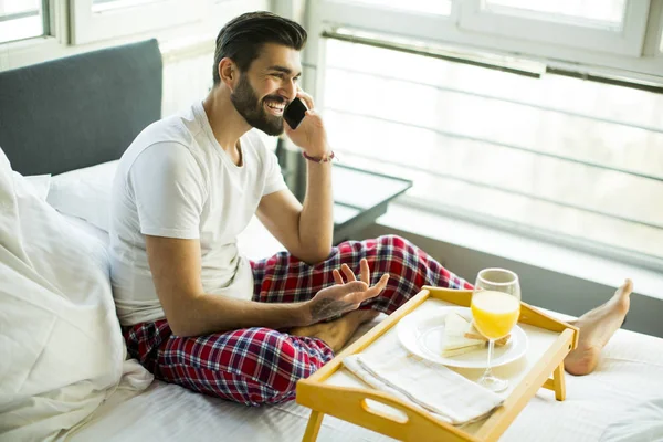 Jeune homme prenant le petit déjeuner au lit et utilisant un téléphone portable — Photo