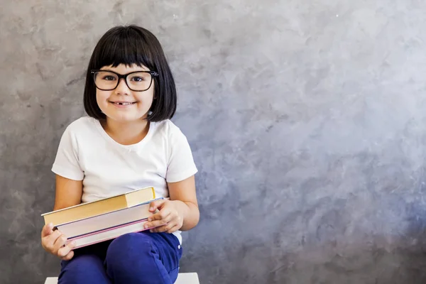 Petite fille aux cheveux noirs avec des lunettes tenant des livres au mur — Photo