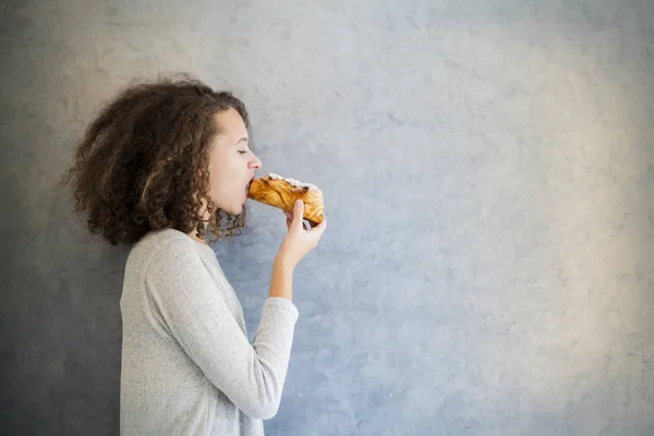 Curar chica pelo rizado comer croissant contra la pared —  Fotos de Stock