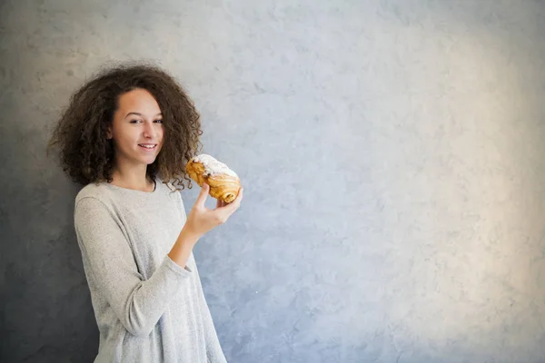 Curar chica pelo rizado comer croissant contra la pared — Foto de Stock