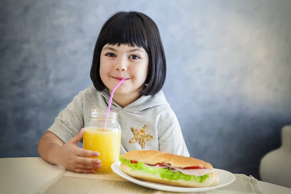 Linda niña de pelo negro bebiendo jugo de naranja y comiendo san — Foto de Stock