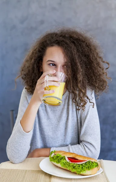 Cabelo encaracolado menina adolescente beber suco de laranja e comer sanduíche — Fotografia de Stock