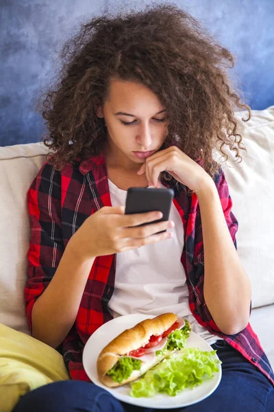 Σγουρά μαλλιά κορίτσι έφηβος χρησιμοποιώντας mibile το τηλέφωνο και τρώνε σάντουιτς — Φωτογραφία Αρχείου