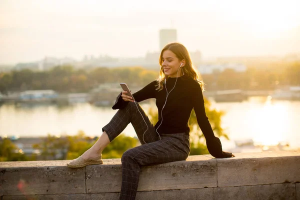 Mujer joven escuchando música desde el teléfono inteligente al aire libre al atardecer — Foto de Stock