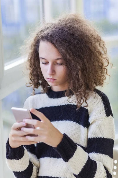 Teen dziewczyna kręcone włosy z telefonu komórkowego przez okno — Zdjęcie stockowe