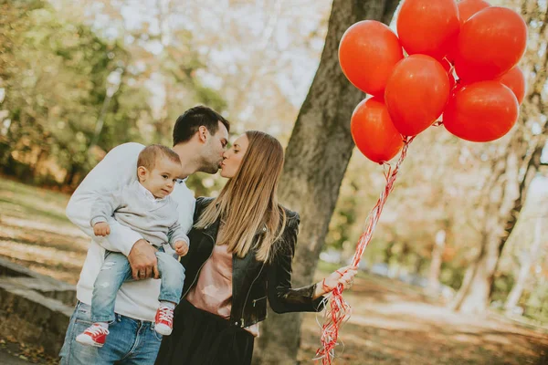 Happy young parents have fun with baby boy in autumn park holding red balloons