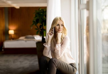 Odanın penceresinde cep telefonuyla ile genç ve güzel sarışın kadın