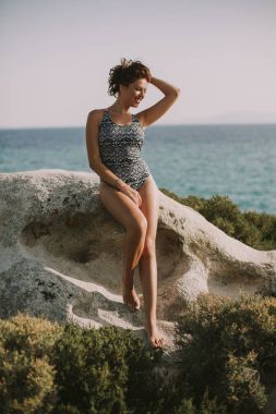 Denizin kenarında kayalık kıyısında güneşli yaz gününde oturan genç ve güzel kadın