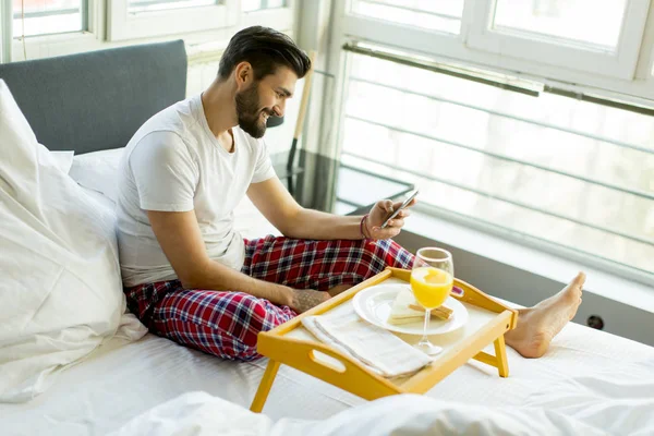 Hombre Joven Desayunando Cama Mientras Usa Teléfono Móvil — Foto de Stock