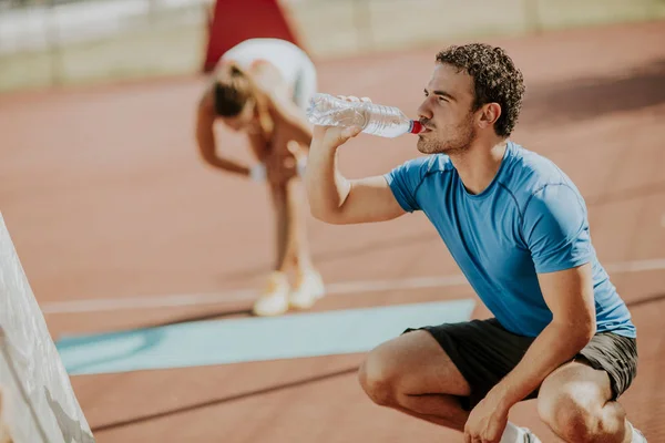 Sportiga Mannen Dricksvatten Medan Ung Kvinna Gör Övning Bakgrunden Utomhus — Stockfoto