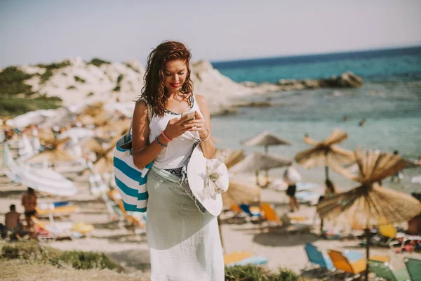 美丽的年轻女子与一个袋子使用手机在海滩上 在阳光明媚的夏日 — 图库照片